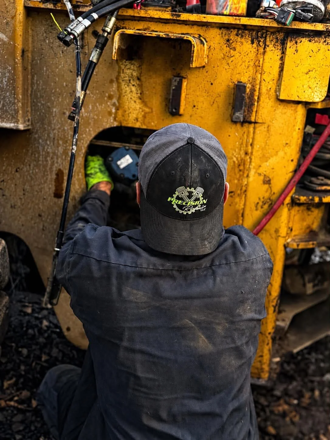 Travis, seen from behind in a Precision Repair hat and dark coveralls, reaching into an access panel on a yellow piece of heavy equipment